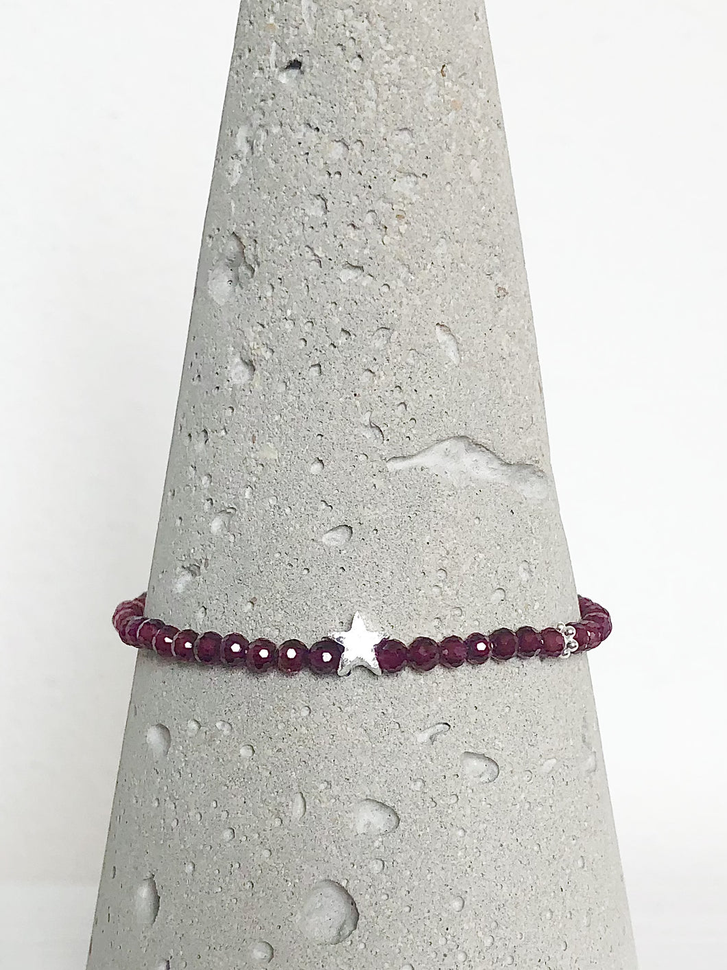 Klassisch-dezentes Armband aus facettiertem rotem Granat und einem mattierten Stern aus Echtsilber, begleitet von einem zierlichen Silberblümchen, Detailansicht auf Betonkegel.