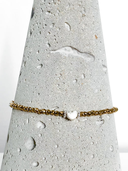 Zierliches Armband aus goldenen Hämatitperlen mit silbernem Herz und einer diamantierten Kugel aus Echtsilber. Detailansicht auf Betonkegel.